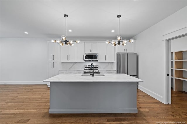 kitchen with an island with sink, light stone counters, a sink, stainless steel appliances, and a notable chandelier