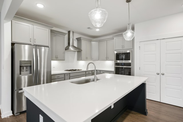 kitchen featuring sink, stainless steel appliances, wall chimney range hood, an island with sink, and pendant lighting