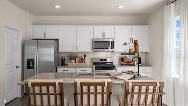 kitchen with a kitchen island with sink, white cabinets, appliances with stainless steel finishes, tasteful backsplash, and light stone counters