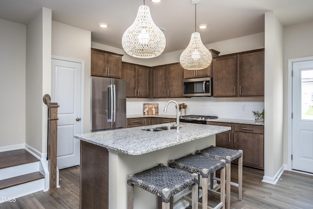 kitchen with light stone countertops, appliances with stainless steel finishes, a kitchen island with sink, sink, and hanging light fixtures