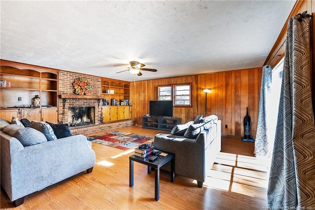 living room featuring a brick fireplace, built in shelves, a textured ceiling, ceiling fan, and light hardwood / wood-style floors