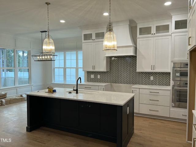 kitchen with white cabinetry, hanging light fixtures, premium range hood, an island with sink, and light hardwood / wood-style floors