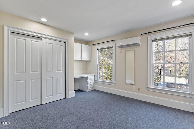 unfurnished bedroom featuring multiple windows, a wall mounted AC, built in desk, and dark colored carpet