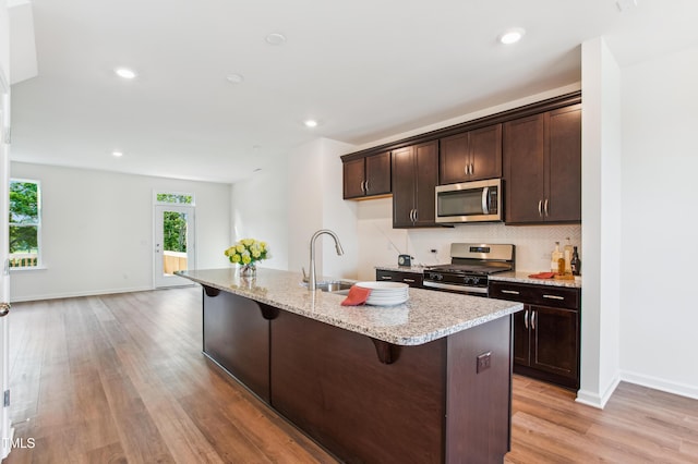 kitchen with dark brown cabinets, light stone counters, an island with sink, and appliances with stainless steel finishes