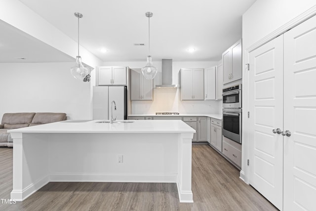 kitchen featuring appliances with stainless steel finishes, a center island with sink, hanging light fixtures, and wall chimney range hood