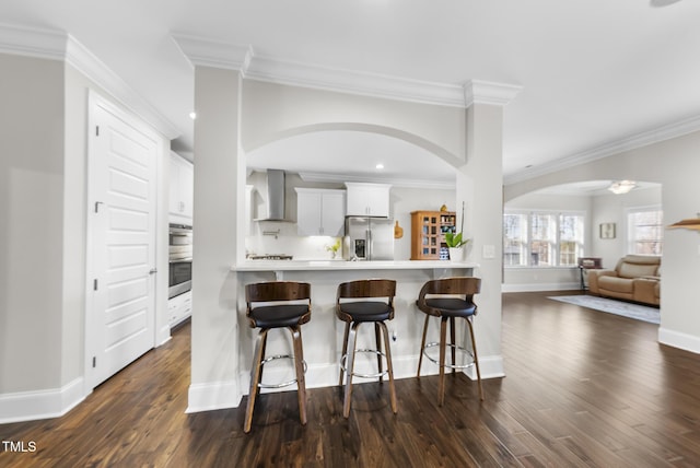 kitchen with white cabinets, appliances with stainless steel finishes, wall chimney exhaust hood, dark wood-type flooring, and a kitchen breakfast bar