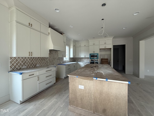 kitchen featuring custom exhaust hood, tasteful backsplash, light wood-type flooring, stainless steel microwave, and a kitchen island with sink