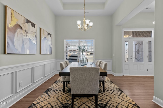 dining space featuring dark wood-style floors, a decorative wall, a raised ceiling, and an inviting chandelier