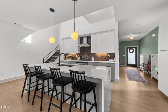 kitchen featuring sink, wall chimney range hood, white cabinetry, an island with sink, and decorative light fixtures