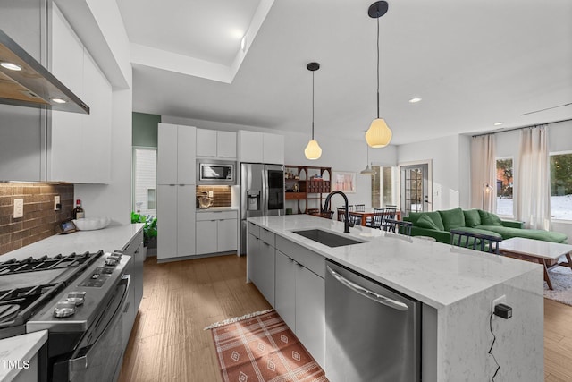 kitchen featuring wall chimney exhaust hood, appliances with stainless steel finishes, white cabinets, a kitchen island with sink, and backsplash