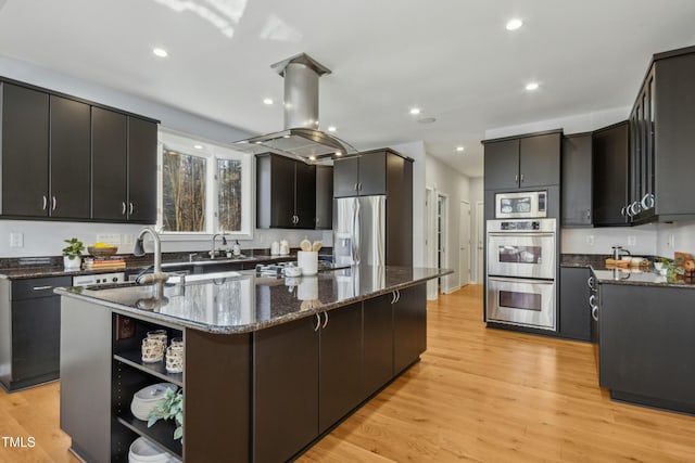 kitchen featuring island exhaust hood, a kitchen island with sink, appliances with stainless steel finishes, light wood-type flooring, and dark stone counters