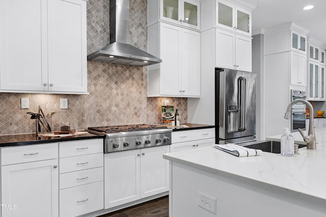 kitchen featuring stainless steel appliances, dark stone counters, white cabinets, wall chimney range hood, and backsplash