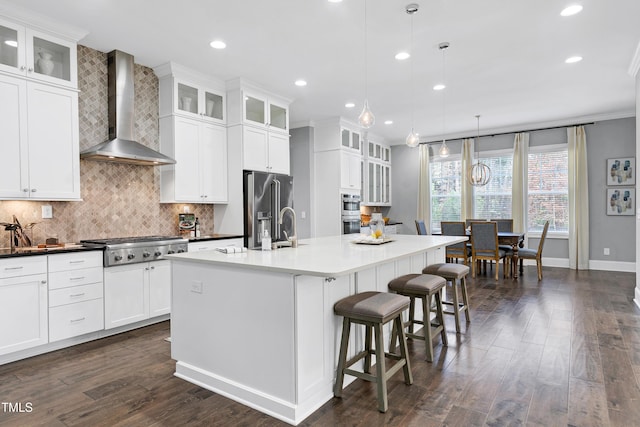 kitchen with white cabinetry, stainless steel appliances, wall chimney range hood, decorative light fixtures, and a kitchen island with sink