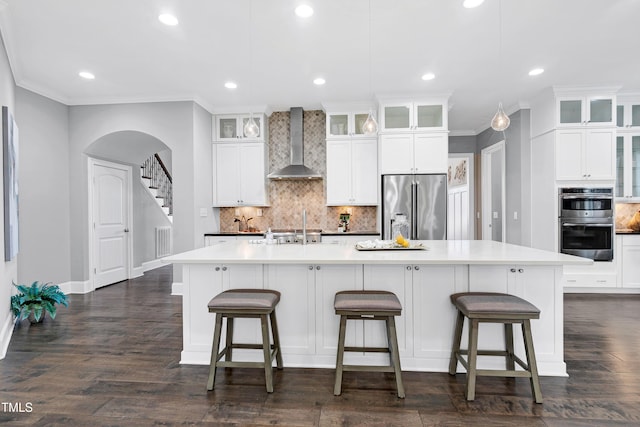 kitchen with white cabinetry, wall chimney exhaust hood, stainless steel appliances, tasteful backsplash, and a center island with sink