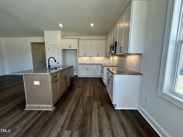 kitchen featuring stainless steel microwave, a kitchen island with sink, white cabinets, a sink, and dark stone counters