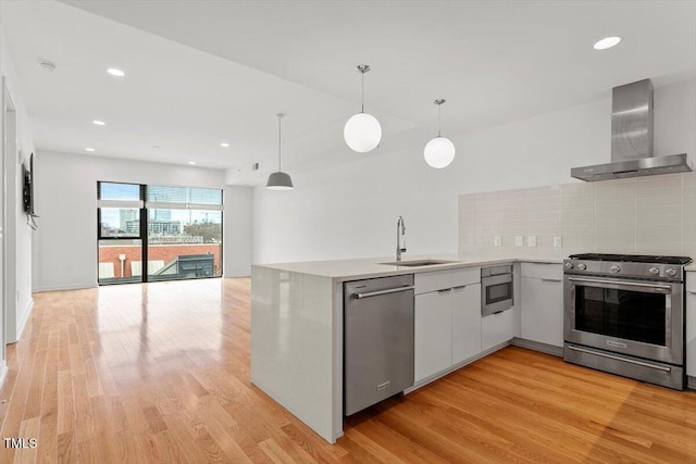 kitchen featuring light wood finished floors, a peninsula, stainless steel appliances, wall chimney range hood, and a sink