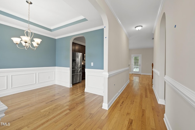 hallway with a chandelier, light wood-type flooring, ornamental molding, arched walkways, and a raised ceiling