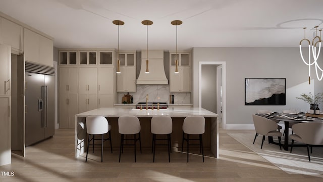 kitchen with pendant lighting, stainless steel built in refrigerator, custom exhaust hood, backsplash, and an island with sink