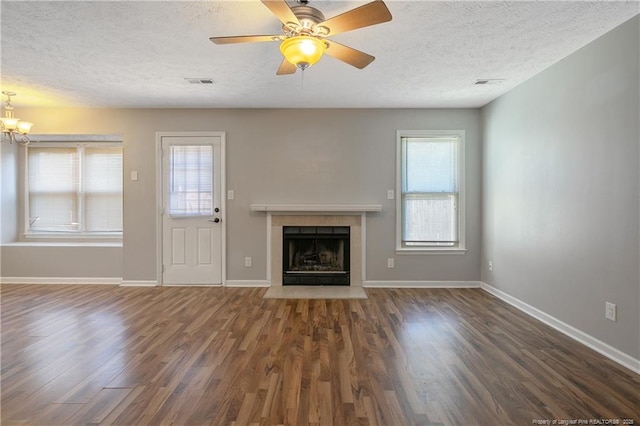 unfurnished living room with ceiling fan with notable chandelier, a tiled fireplace, a textured ceiling, and dark wood-type flooring