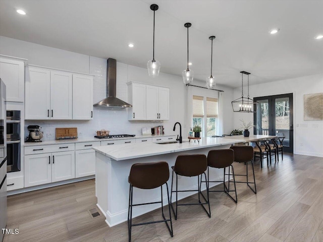 kitchen with white cabinets, a center island with sink, wall chimney range hood, and hanging light fixtures