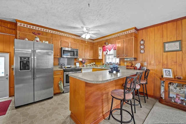 kitchen featuring kitchen peninsula, stainless steel appliances, wood walls, a kitchen bar, and ceiling fan