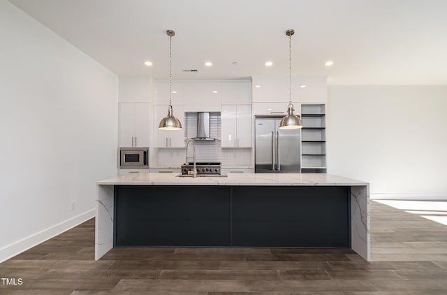 kitchen with built in appliances, white cabinetry, wall chimney exhaust hood, a large island with sink, and light stone countertops