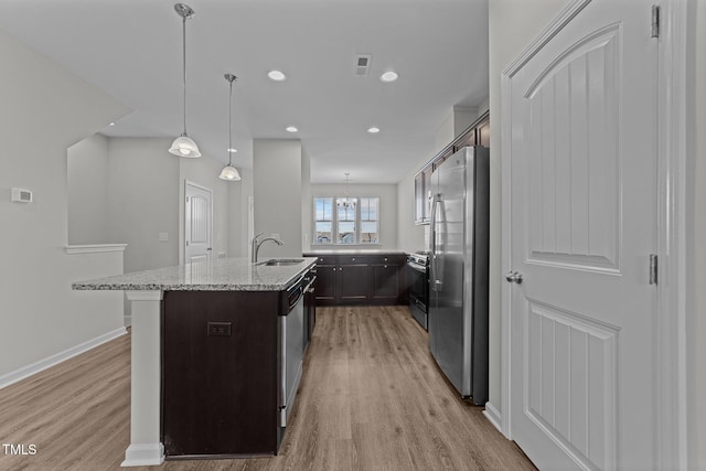 kitchen featuring light stone counters, an island with sink, dark brown cabinets, pendant lighting, and appliances with stainless steel finishes