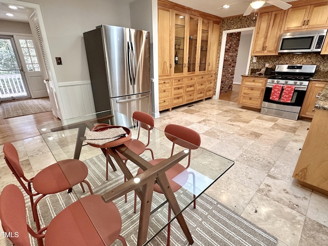 kitchen featuring a ceiling fan, wainscoting, glass insert cabinets, stainless steel appliances, and backsplash