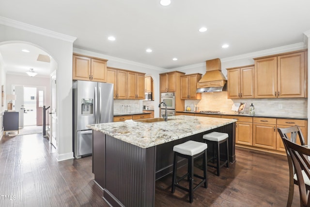 kitchen with premium range hood, a center island with sink, a sink, stainless steel appliances, and arched walkways