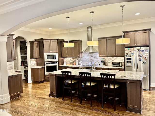 kitchen with tasteful backsplash, wall chimney range hood, a center island with sink, pendant lighting, and appliances with stainless steel finishes