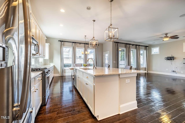 kitchen with appliances with stainless steel finishes, white cabinets, hanging light fixtures, light stone counters, and a center island with sink