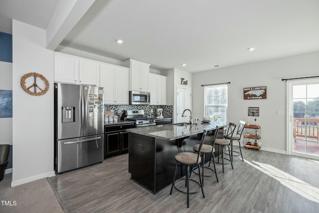 kitchen with a kitchen breakfast bar, dark stone countertops, stainless steel appliances, an island with sink, and white cabinetry