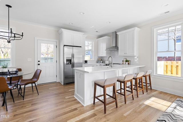 kitchen with white cabinets, wall chimney exhaust hood, kitchen peninsula, stainless steel fridge with ice dispenser, and pendant lighting