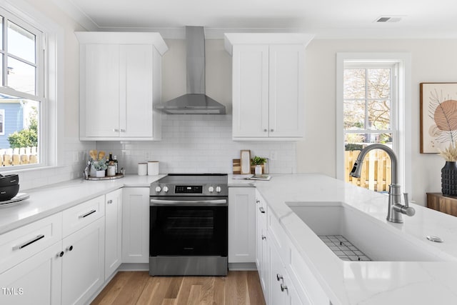 kitchen with stainless steel electric stove, wall chimney exhaust hood, sink, white cabinetry, and tasteful backsplash