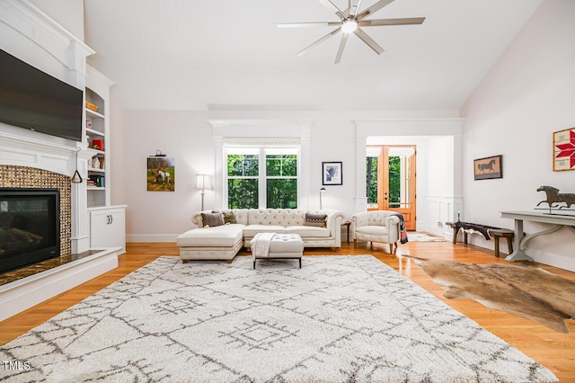 living room featuring ceiling fan, high vaulted ceiling, light hardwood / wood-style floors, a tiled fireplace, and built in shelves