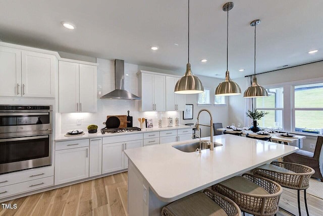 kitchen featuring wall chimney range hood, pendant lighting, sink, an island with sink, and white cabinets