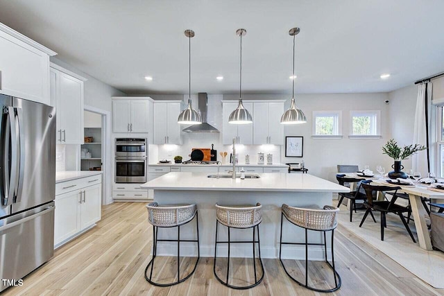 kitchen with stainless steel appliances, a kitchen island with sink, hanging light fixtures, wall chimney exhaust hood, and white cabinets