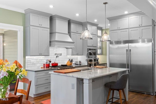 kitchen featuring built in refrigerator, a kitchen breakfast bar, custom range hood, light stone countertops, and a kitchen island with sink