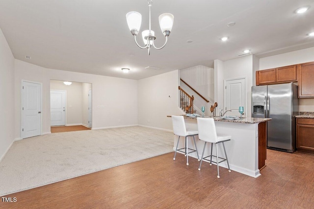 kitchen featuring an inviting chandelier, stainless steel fridge, a kitchen island with sink, light stone countertops, and pendant lighting