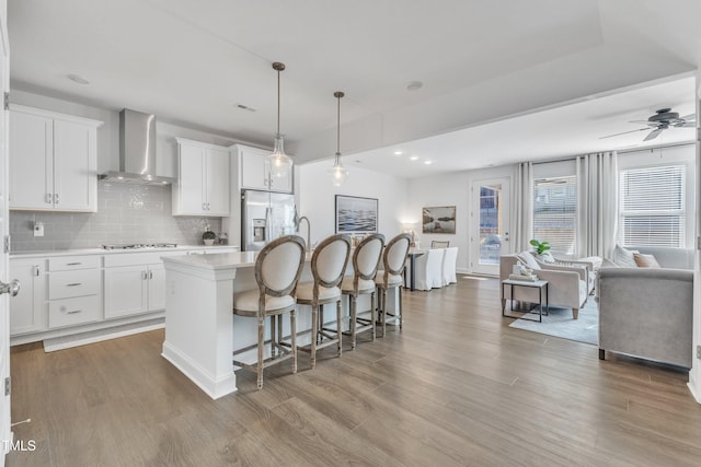 kitchen with an island with sink, stainless steel refrigerator with ice dispenser, pendant lighting, wall chimney exhaust hood, and white cabinets