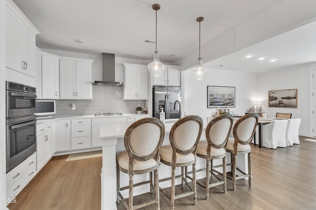 kitchen featuring white cabinetry, stainless steel appliances, a kitchen island with sink, decorative light fixtures, and wall chimney exhaust hood