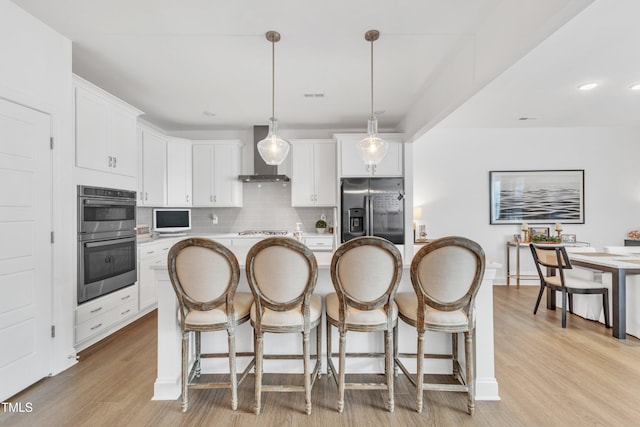 kitchen with decorative light fixtures, wall chimney range hood, a kitchen island, appliances with stainless steel finishes, and white cabinets