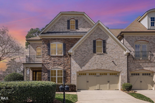 french country inspired facade with a balcony, driveway, an attached garage, and brick siding