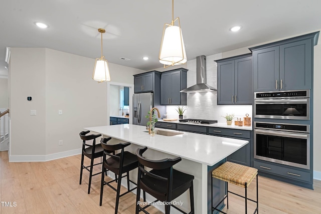 kitchen with a center island with sink, stainless steel appliances, light countertops, wall chimney range hood, and a sink