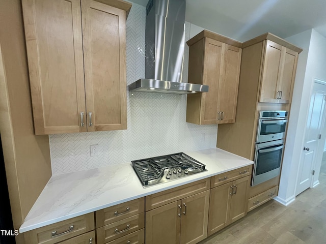 kitchen with wall chimney range hood, stainless steel appliances, decorative backsplash, light stone counters, and light hardwood / wood-style flooring