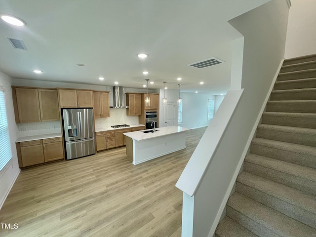 kitchen with wall chimney exhaust hood, stainless steel appliances, tasteful backsplash, light hardwood / wood-style flooring, and a center island with sink