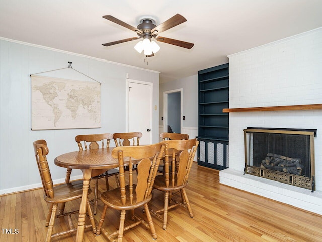 dining room featuring ceiling fan, a brick fireplace, built in features, ornamental molding, and light hardwood / wood-style flooring