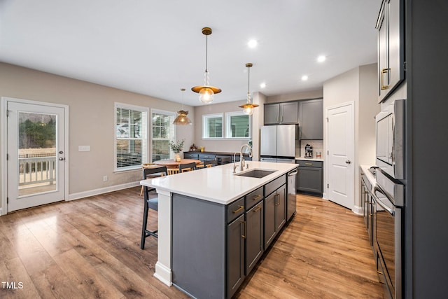 kitchen with appliances with stainless steel finishes, hanging light fixtures, sink, light wood-type flooring, and an island with sink