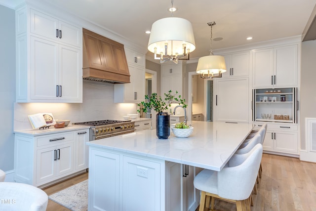 kitchen featuring hanging light fixtures, stainless steel stove, an island with sink, custom range hood, and white cabinets