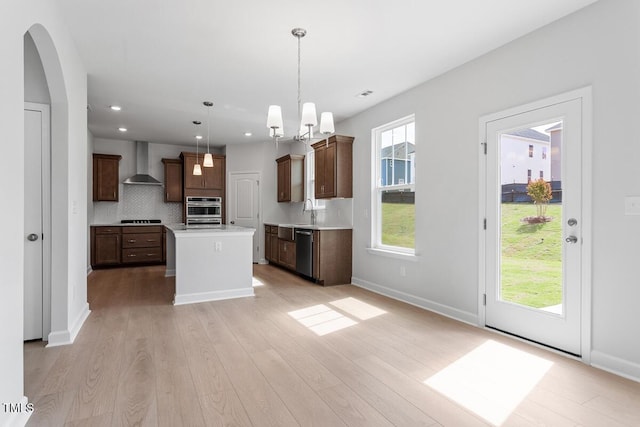 kitchen featuring light wood finished floors, light countertops, wall chimney exhaust hood, decorative backsplash, and appliances with stainless steel finishes